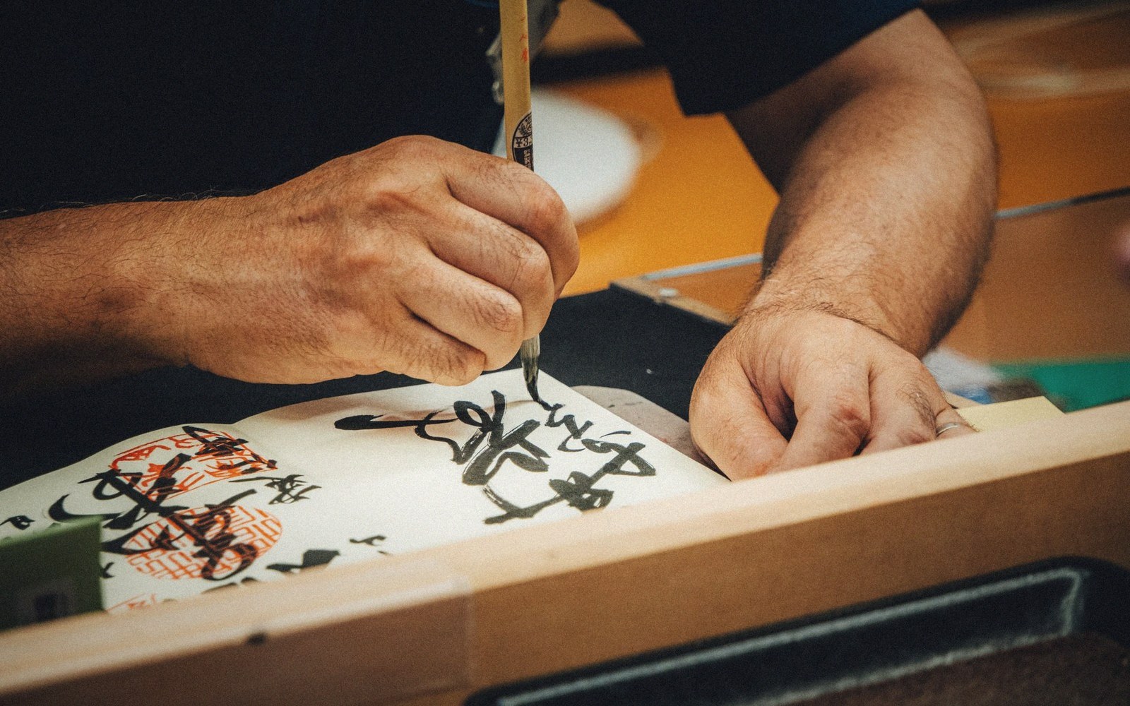 Hands using a brush to write black calligraphy characters on paper with red stamps