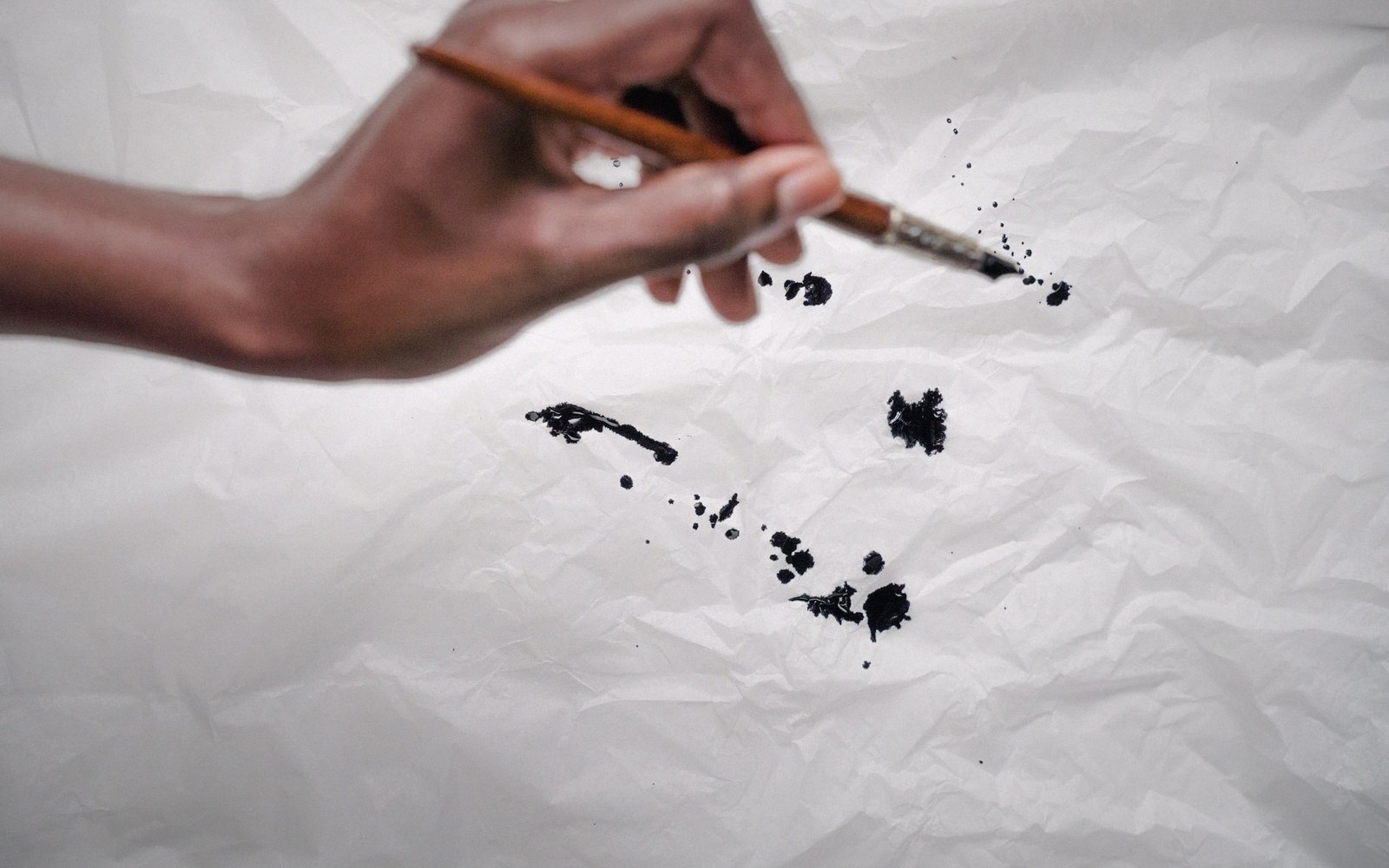 A hand holds a paintbrush flicking black ink splatters onto crumpled white paper